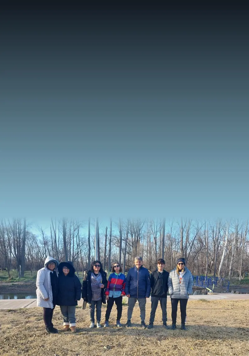 Grupo de sete pessoas em fila num parque em Neuquén, Argentina, com árvores e um riacho ao fundo